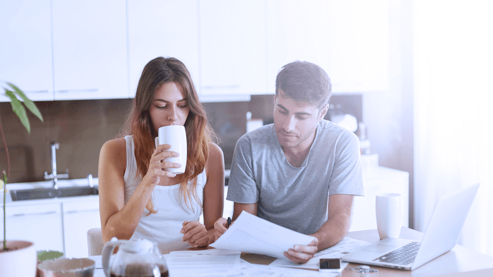 A woman drinks from a mug while a man looks at papers at a kitchen table. They appear focused, with documents, a laptop, and other items spread out in front of them.