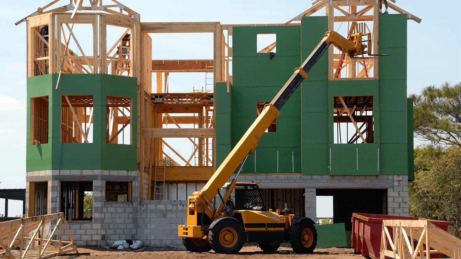 A yellow telescopic forklift lifts materials to the upper level of a partially constructed house with exposed wooden framing and green sheathing, surrounded by construction debris and a red dumpster.