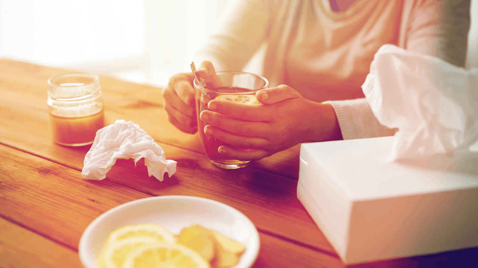 A person holding a mug at a wooden table with tissues, a plate of lemon slices, and a tissue box, suggesting they may be feeling unwell or soothing a cold.