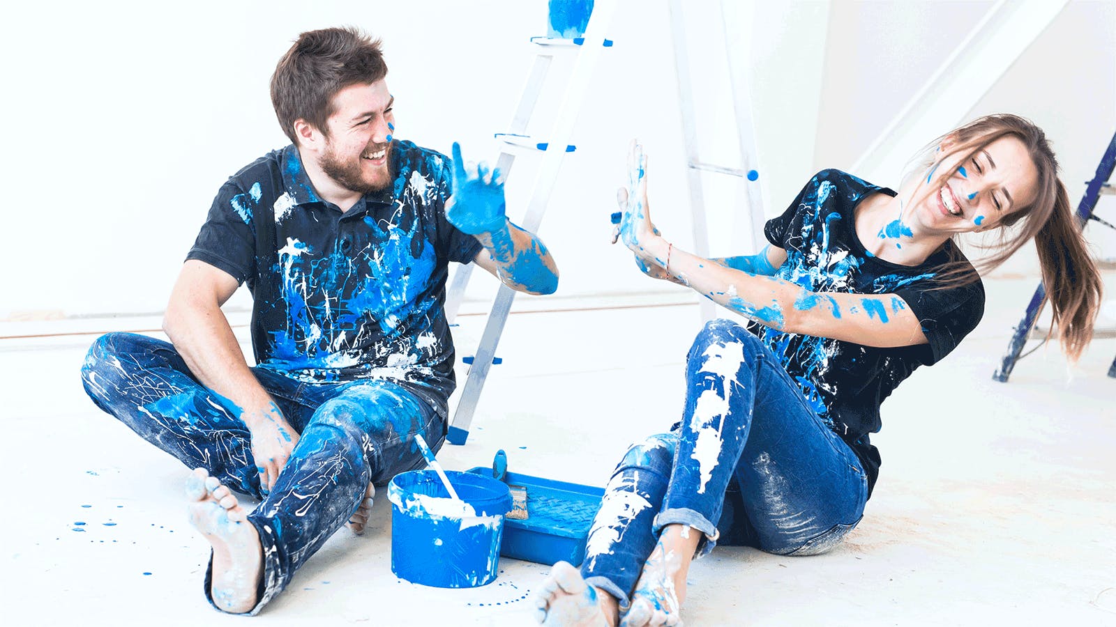 A man and woman sit on the floor, playfully covered in blue paint, smiling and laughing together. Paint cans and a ladder are nearby, suggesting they are painting a room.