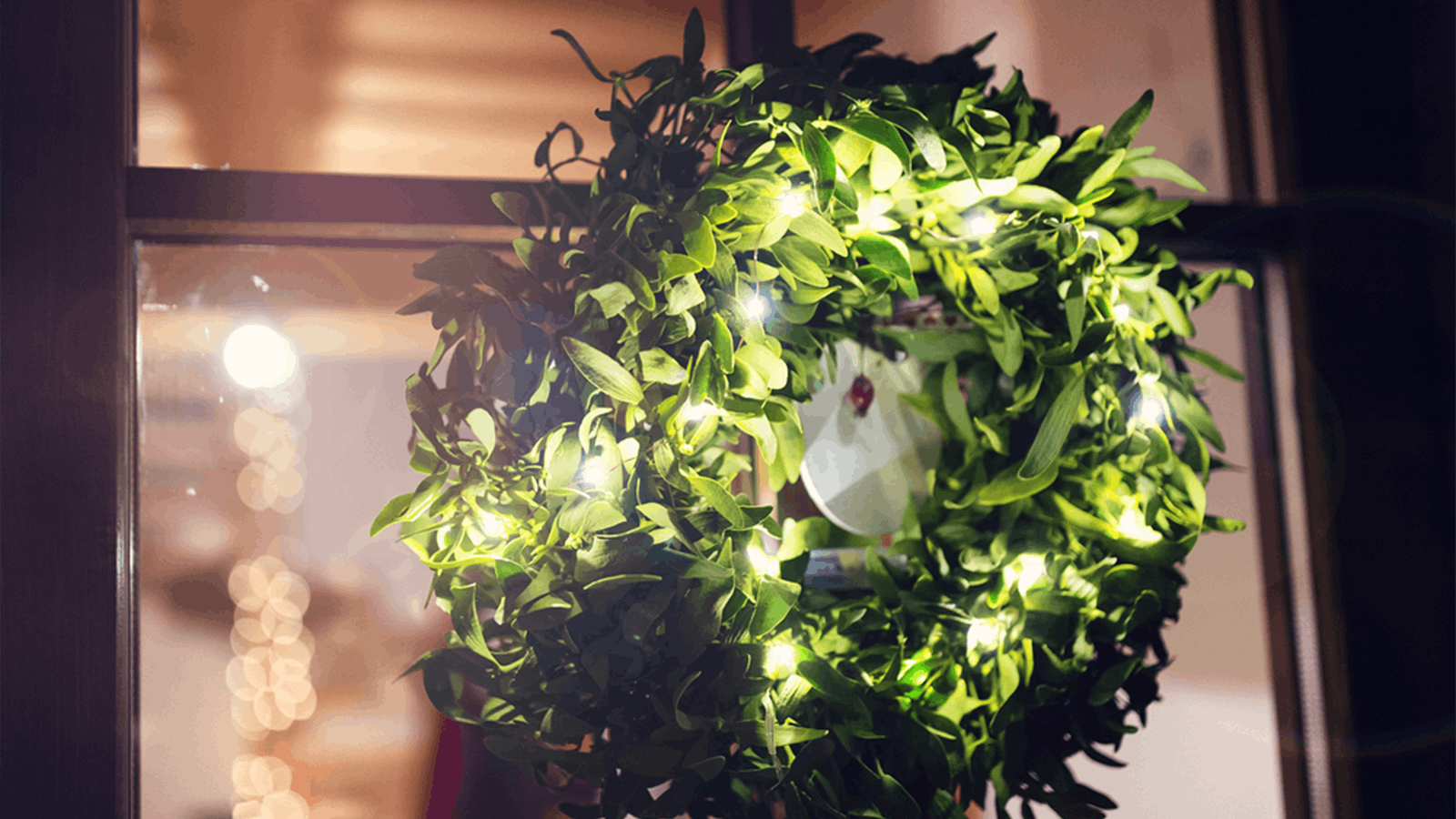 A green holiday wreath decorated with small white string lights hangs on a window, with warm indoor lights softly glowing in the background.