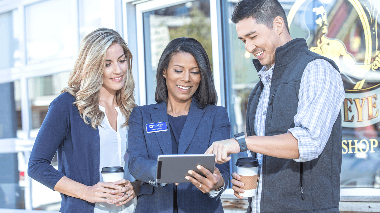 Three people stand outside, smiling and holding coffee cups, while looking at a tablet held by the woman in the center, who is wearing a navy blazer with a nametag.