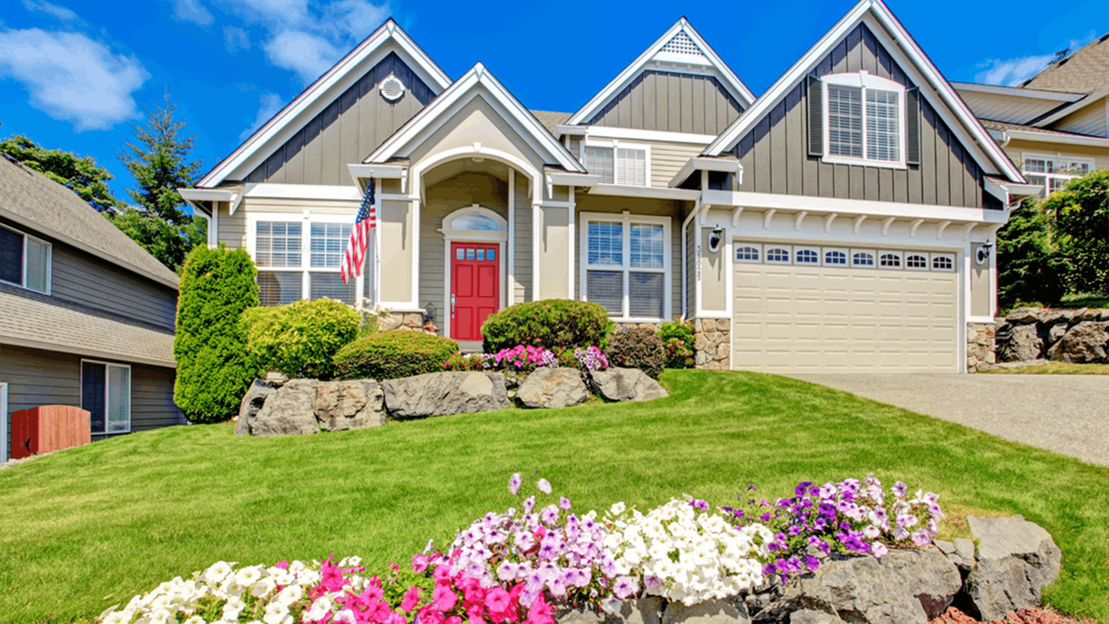 A two-story suburban house with gray siding, a red front door, large windows, and an American flag. The front yard features manicured grass, colorful flowers, bushes, and a stone-bordered driveway.