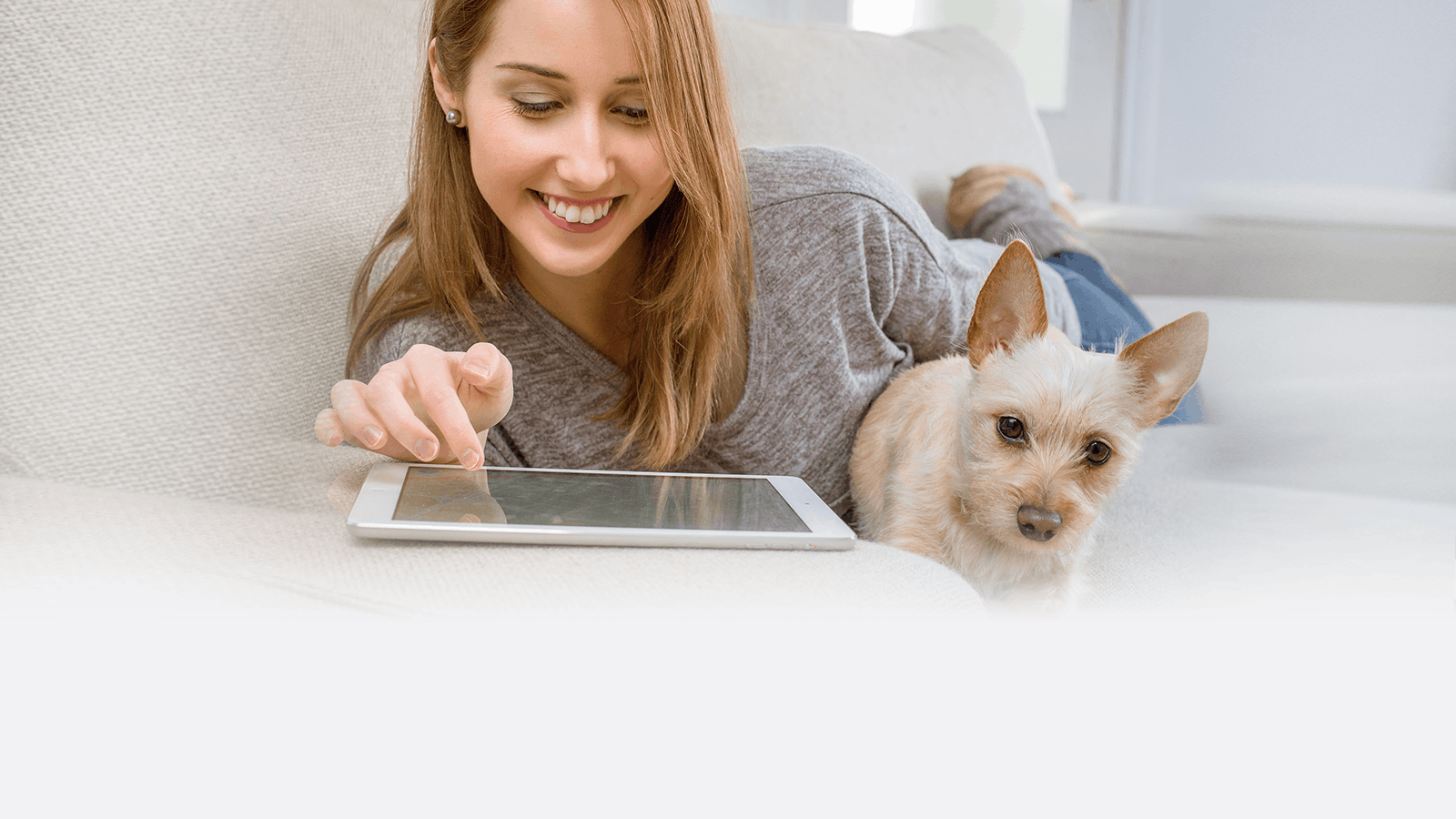 A woman lying on a sofa smiles while using a tablet, with a small, light brown dog resting beside her.