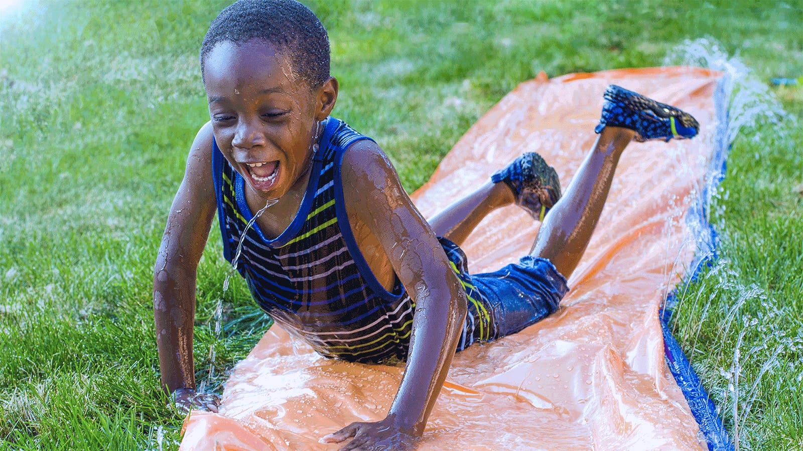 A smiling child in a striped tank top and shorts slides belly-down on a wet slip-and-slide outdoors, surrounded by green grass and splashing water.