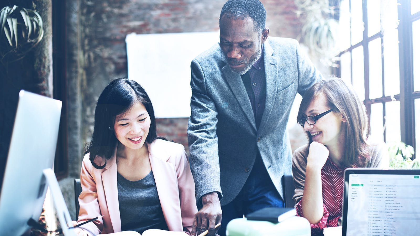 Three colleagues work together in a bright office. A man stands and points at a document while two women sit at the desk, smiling and looking at the paper. Computers and office supplies are on the table.