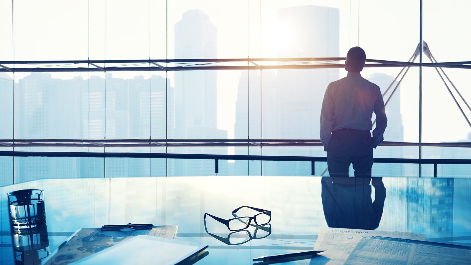 A person in business attire stands by large office windows overlooking a city skyline, with sunlight streaming in. On the glass desk in the foreground are papers, a pen, a glass of water, and eyeglasses.