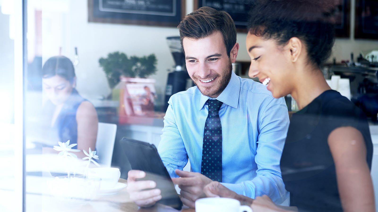 Two people sit at a table in a bright cafe, smiling and looking at a tablet together. A third person works in the background. The scene appears friendly and collaborative.