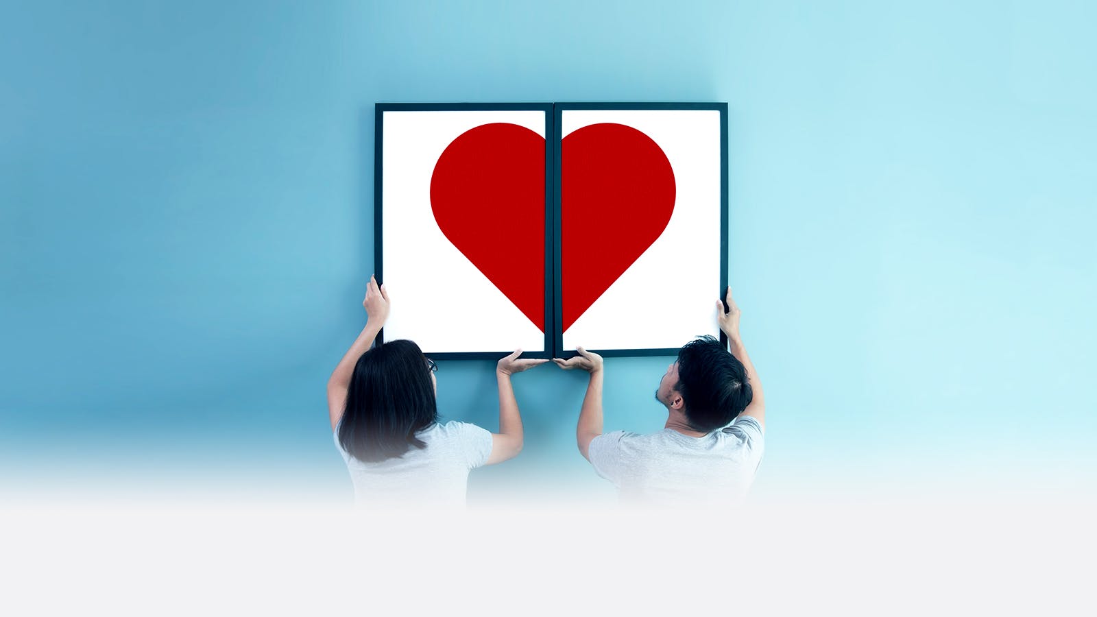 A woman and a man hold up two framed halves of a red heart side by side, forming a complete heart against a blue background.