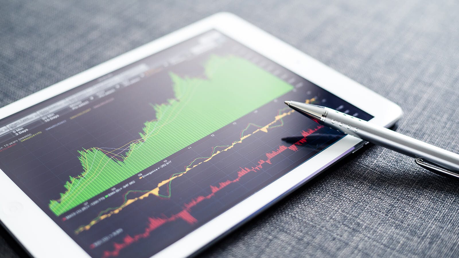 A tablet displaying financial graphs with green, yellow, and red lines showing stock market data. A silver pen rests on the screen, and the background is a textured grey surface.