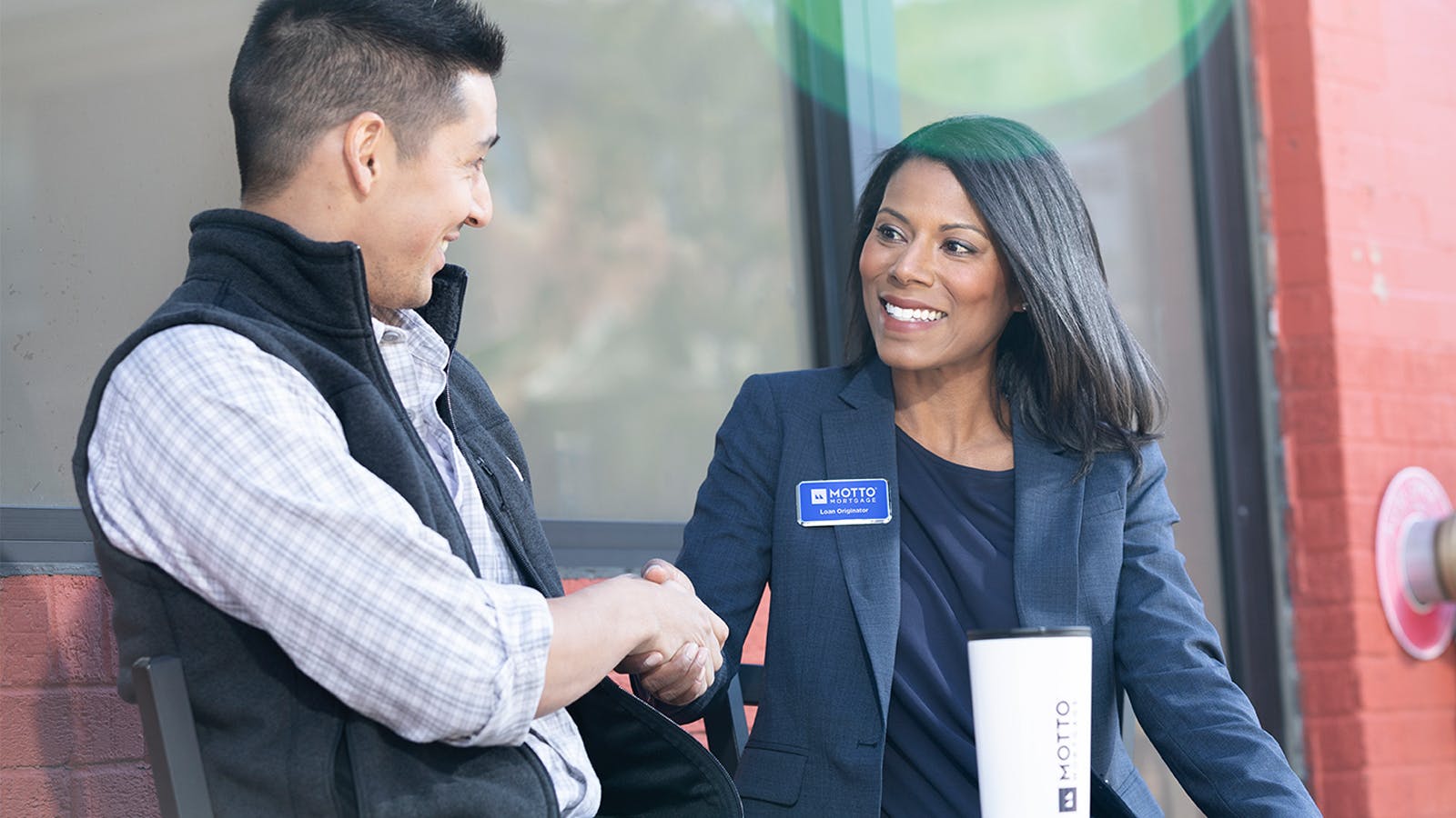 A woman in business attire with a name badge smiles and shakes hands with a man outdoors. Both appear friendly and engaged. A travel mug with the Motto logo sits on the table between them.
