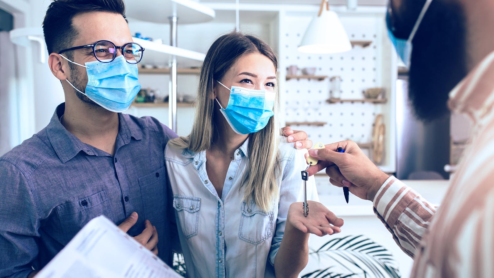A smiling couple wearing masks stands indoors as a person hands them a set of keys, suggesting they are receiving keys to a new home. The woman holds out her hand to accept the keys.