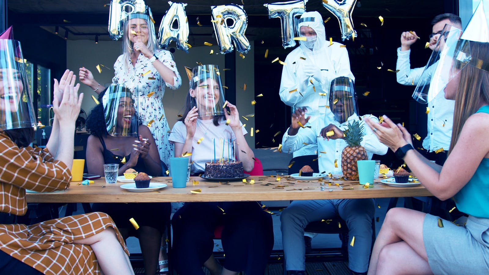 A group of people wearing party hats and face shields celebrate at a table with cake, snacks, drinks, and a pineapple. Silver balloons spell “PARTY” in the background. Confetti fills the air as everyone claps and cheers.