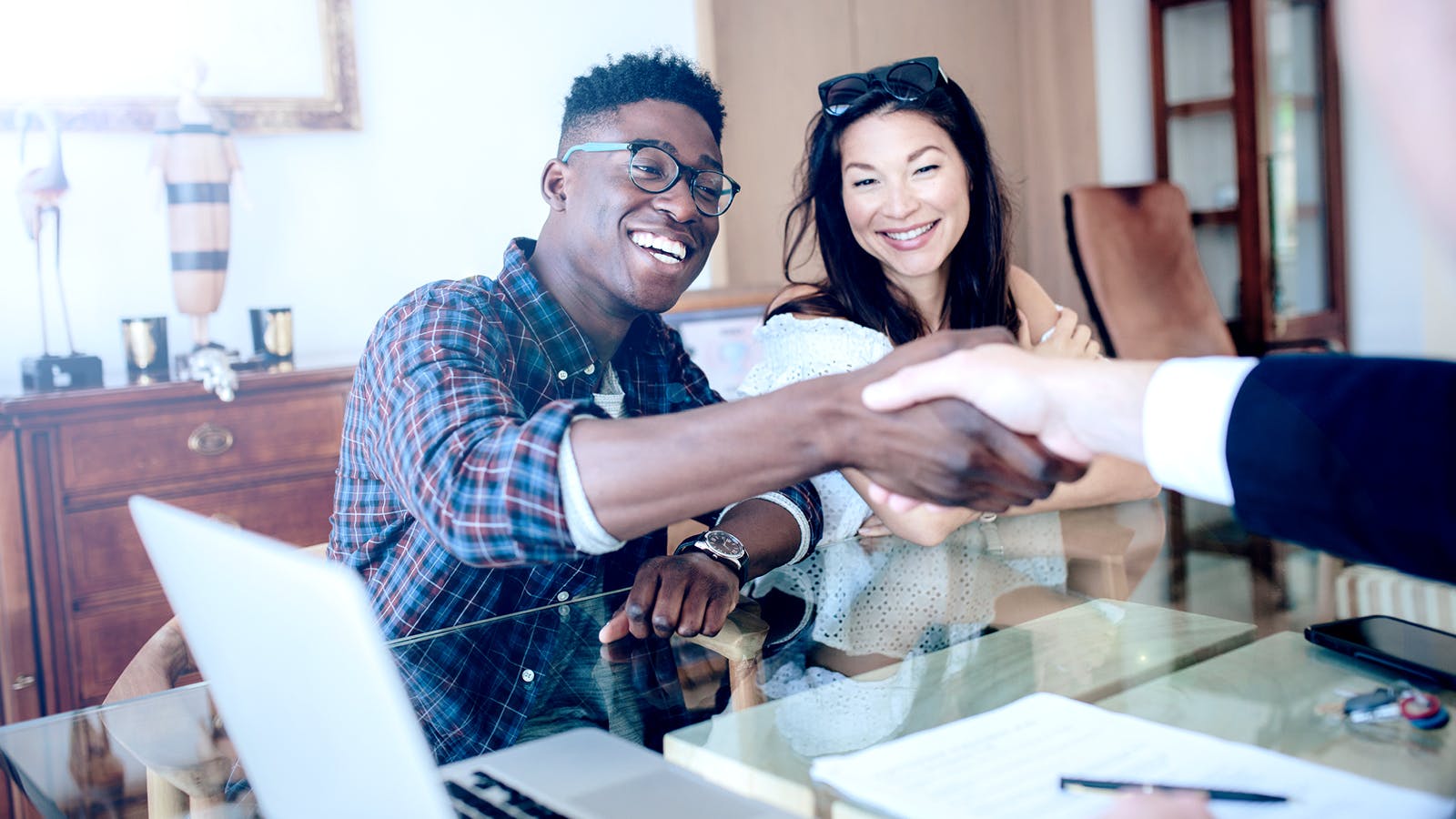 A smiling man and woman sit at a desk; the man is shaking hands with another person across the table, suggesting a successful business meeting. A laptop and documents are on the glass-topped desk.