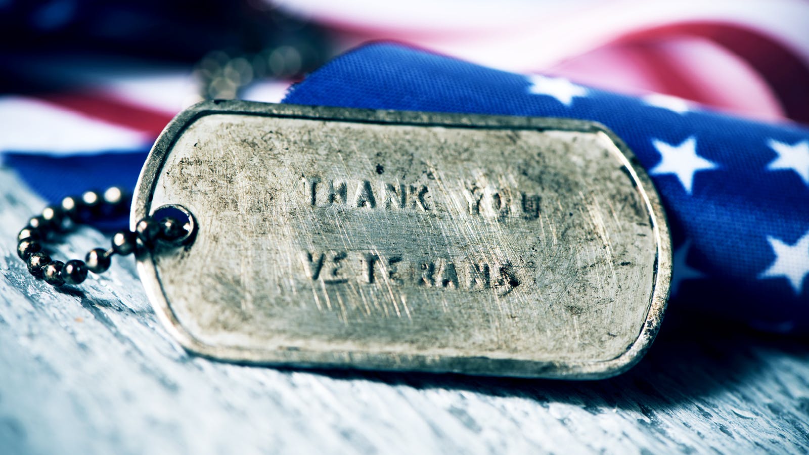 A close-up of a military dog tag engraved with Thank You Veterans rests on a wooden surface, with an American flag featuring white stars on a blue background in the blurred background.