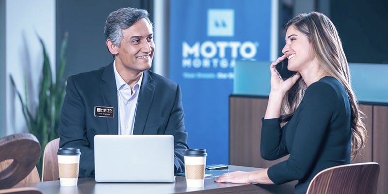 Photo of a mortgage broker meeting with a real estate agent. The agent is speaking on the phone with a homebuyer and the broker is adding information to the computer.