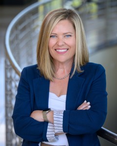 Gill_Kelly_WEB-4341-3 copy A woman with blonde hair wearing a blue blazer and white top stands with arms crossed, smiling. She is in front of a curved metal railing in a bright indoor setting.